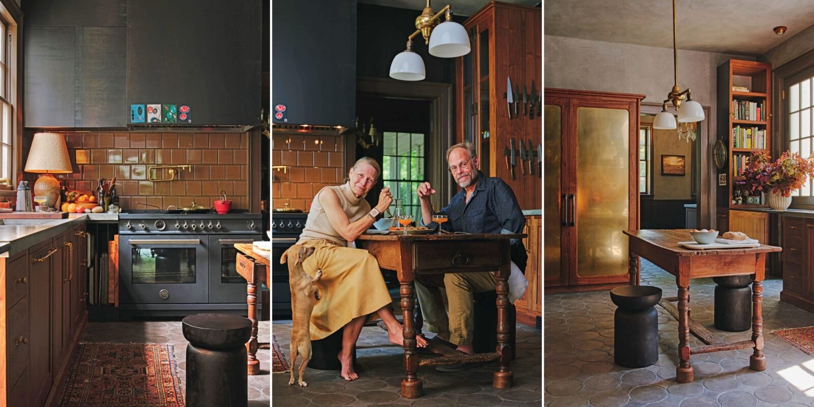 A kitchen with brown tile and dark grey cabinets and stoves; a couple sits at a table with a dog at their feet; an antique farm table under milky lights