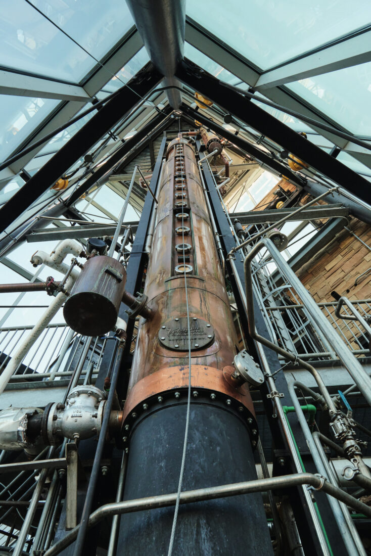 Looking up at a soaring copper still in an industrial building.