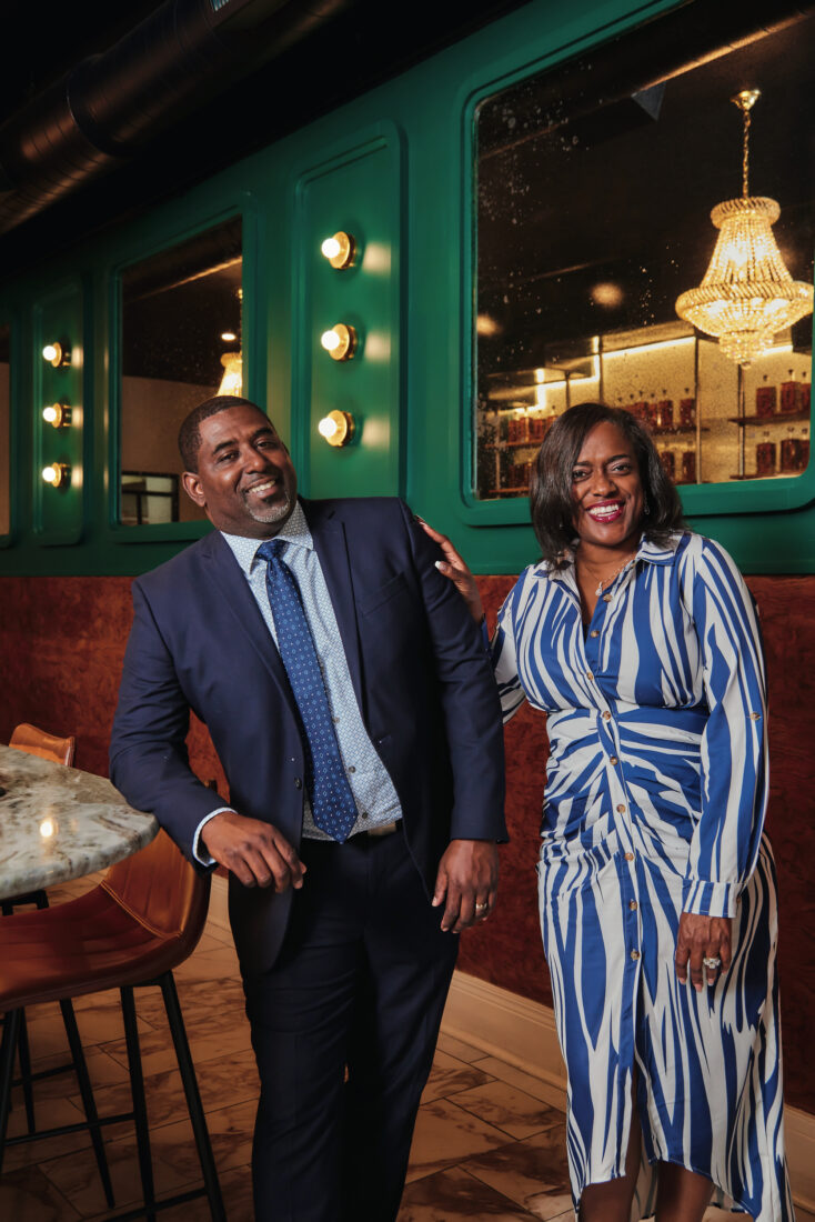 A man wearing a blue jacket and a woman wearing a white and blue stripped dress smile in a bar against a dark green wall