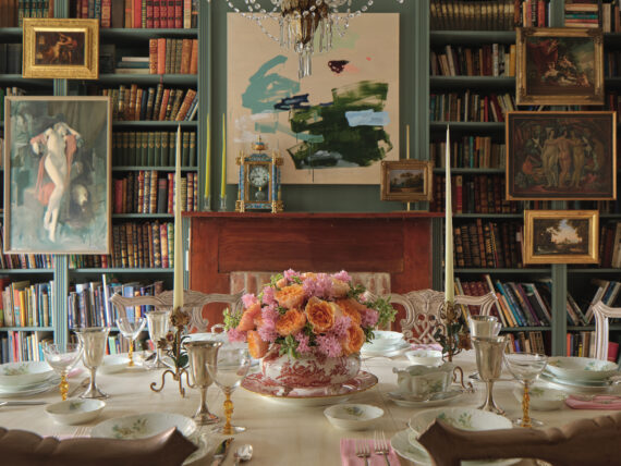 A dining room with dark sage green bookshelves, paiuntings, a crystal chandelier, and a round set table with a bouquet