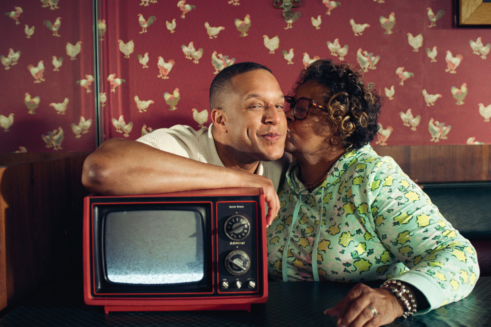 A portrait of Craig Melvin and his mother. They sit in a booth of a restaurant with chicken-patterned wallpaper. She kisses his cheek and his arm is draped over an old television