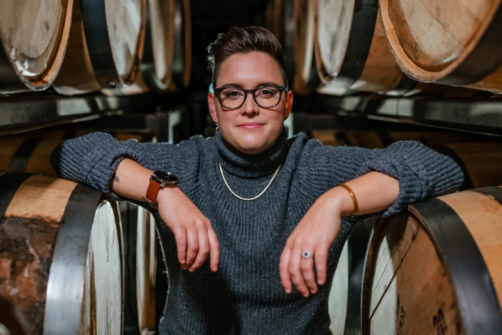 A portrait of a woman wearing a grey sweater and glasses between wooden casks