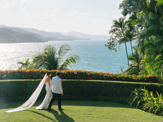 A bride and groom walk across a lawn with palm trees and water in the distance